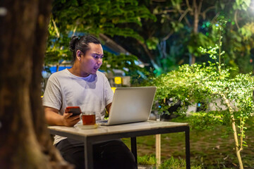 Young Asian man, digital nomad working remotely from cafe, using laptop and smartphone. young Asian men are hanging out in a cafe with a nature theme at night