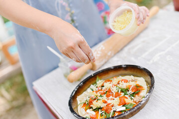 Close-up of hands preparing pizza with cheese and fresh ingredients outdoors. Ideal for summer gatherings, picnics, and casual cooking activities.