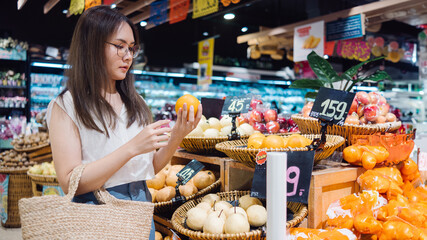 A woman with a woven bag is selecting fresh oranges in a brightly lit supermarket's produce section. Ideal for illustrating grocery shopping, healthy eating, or retail environments.