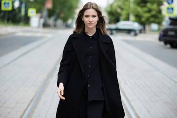 Attractive caucasian young woman in black coat walking down city street on warm autumn day. Outdoors portrait. Model in stylish clothes. Lifestyle, fashion concept.