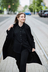 Obraz premium Attractive caucasian young woman in black coat walking down city street on warm autumn day. Outdoors portrait. Model in stylish clothes. Lifestyle, fashion concept.