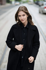Obraz premium Attractive caucasian young woman in black coat walking down city street on warm autumn day. Outdoors portrait. Model in stylish clothes. Lifestyle, fashion concept.