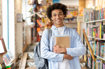 Diligent black male student standing in university library, surrounded by bookshelves, engaged in...