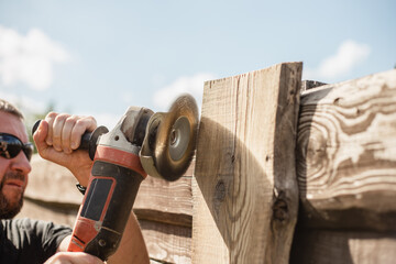 A man with a powerful tool with an angle grinder cleans the wood of the fence - grinding the boards with a coarse grinder brush