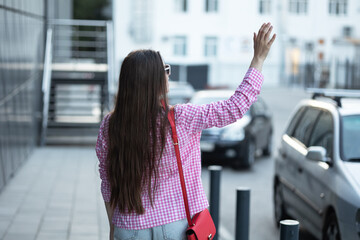 woman pink and white checkered shirt hails taxi city street. Standing on the sidewalk red purse slung over her shoulder and looking back approaching car. raised hand up. hand gesture. vichy cage