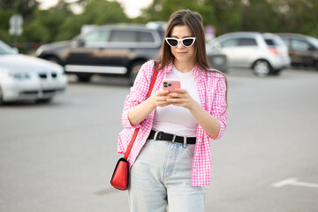  woman uses technology of her smartphone on the street. artificial intelligence. send a voice message. online social networks. woman wearing pink gingham shirt jeans white sneakers standing parking 