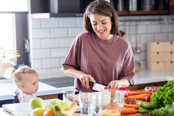 A cheerful mother is making a healthy meal with lots of fruit and vegetables, and her cute little baby daughter is keeping her company