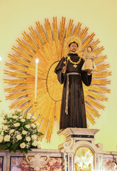 Statue of saint anthony of padua holding baby jesus and a lily, standing inside a church in front of a golden halo