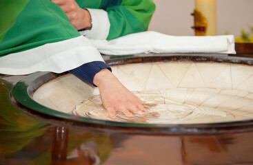 Priest in green liturgical vestments blessing holy water in a marble baptismal font inside a church