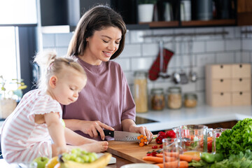 A cheerful mother is making a healthy meal with lots of fruit and vegetables, and her cute little baby daughter is keeping her company