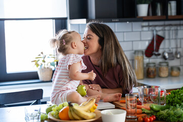 Cheerful mother and her cute little baby are eating healthy fruit and vegetables in the home kitchen