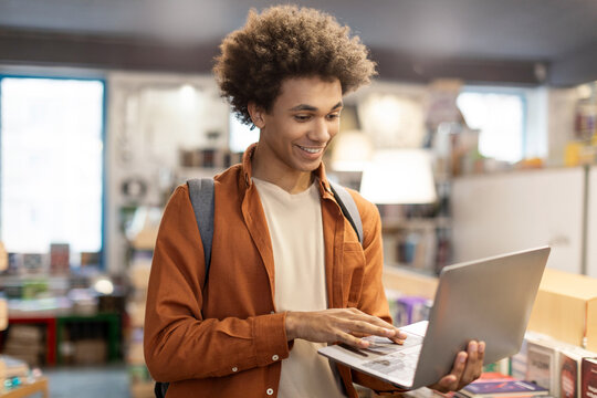 Busy black guy student using laptop standing in university or college library among bookshelves, checking information and smiling