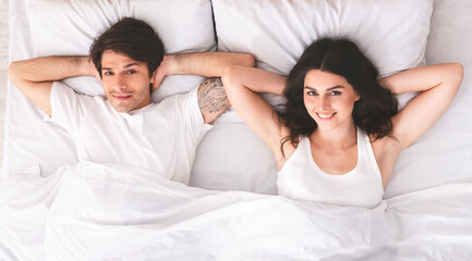 Fototapeta premium A young couple lies in bed together, seemingly after just waking up. They are both smiling and look relaxed and content. They are wearing casual clothes and are lying on white sheets and pillows.