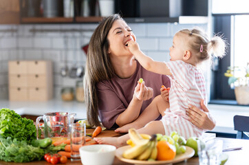 Cheerful mother and her cute little baby are eating healthy fruit and vegetables in the home kitchen