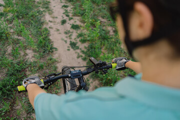 A woman wearing a helmet rides a black mountain bike along a dirt path. The path runs through an open grassy field, with houses and trees visible in the background.