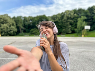 Happy cool woman wearing headphones dancing alone in the park. Young adult happy girl sings holding phone like mic. and hand towards cameral. Healthy lifestyle and happiness concept