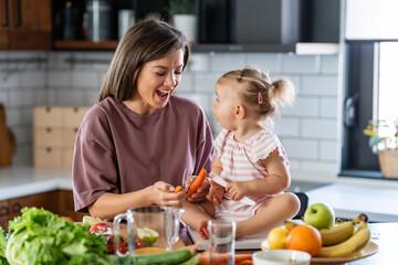 Cheerful mother and her cute little baby are eating healthy fruit and vegetables in the home kitchen