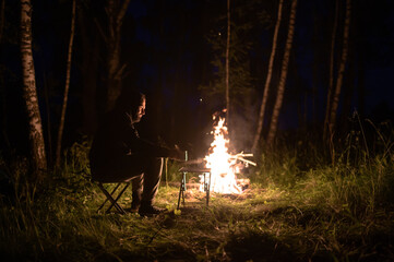 A man sits at night by a campfire in summer.