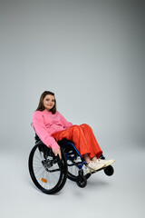 A brunette young woman in a pink sweatshirt and orange pants smiles while sitting in a wheelchair in a studio setting.