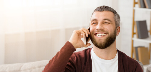 A man sitting comfortably on a couch, engaged in a phone conversation. He is holding a cell phone to his ear, gesturing while speaking. The background is a typical living room setting.