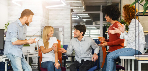 In the office setting, a diverse group of coworkers are seated closely together, engaged in a meeting or discussion, actively converse, gesturing and looking at each other while seated around a table.