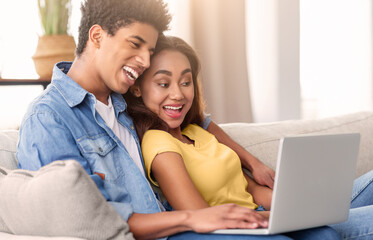 A teen couple is sitting on a couch together, using a laptop and laughing. The guy has his arm around the girl and they both appear to be enjoying each others company.