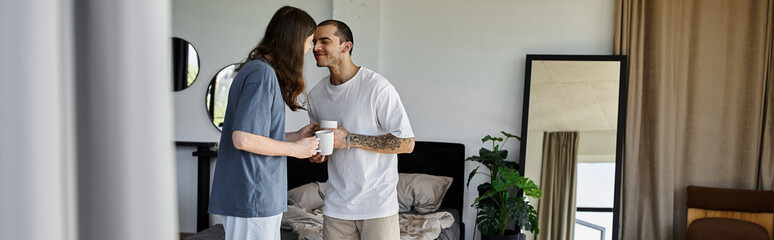 A young gay couple stands in a bedroom, holding mugs and looking lovingly into each others eyes. © Bliss