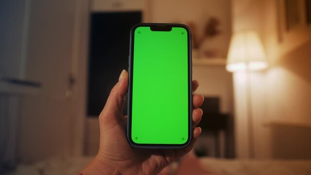 Young girl using smartphone with green screen lying on the bed while chatting with friends, reading ebook, surfing in social media.