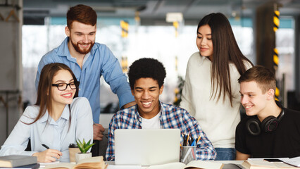 A diverse group of people are gathered around a table indoors. They are actively engaged and focused on a laptop screen, likely discussing work-related matters or collaborating on a project.