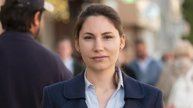 A woman in a formal jacket and shirt stands in the middle of the street and looks at the camera. A crowd of people walks around her. Daily bustle concept