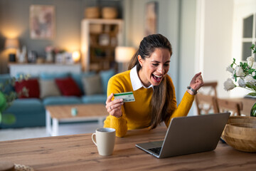 Enthusiastic charming woman feeling excited looking at laptop screen holding credit card, raising fist in yes gesture satisfied with online payment possibilities, rejoicing approved loan.