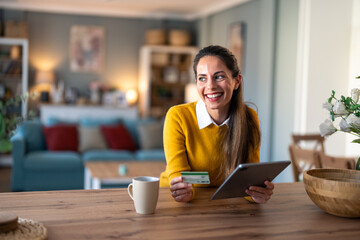 Happy smiling woman entrepreneur holding digital tablet and credit card while sitting at desk at home and looking aside.