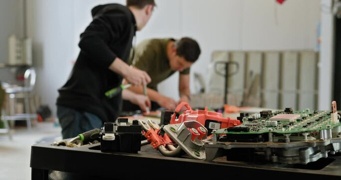 Two technicians working in a repair shop on electric vehicle batteries. Focus on battery components on the table with technicians in the background. Electric car. Pack of battery cells module. EV car