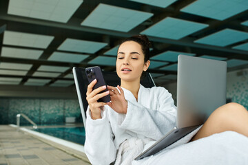 A young, beautiful brunette woman relaxes in a bathrobe by an indoor spa pool, focused on her cell phone.