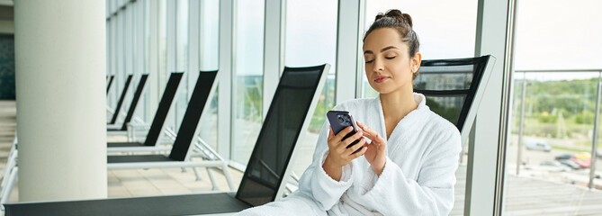 A young brunette woman sitting in a chair, captivated by her cell phone screen, in an indoor spa...