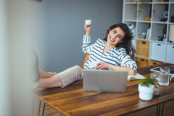 A cheerful Caucasian businesswoman relaxes with her feet up, sipping coffee while multitasking on a...