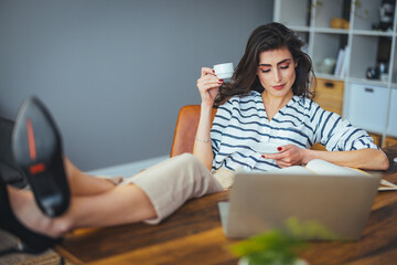 A professional Caucasian woman enjoys a coffee break in her office, her feet up while working on a laptop, highlighting a blend of comfort and productivity.