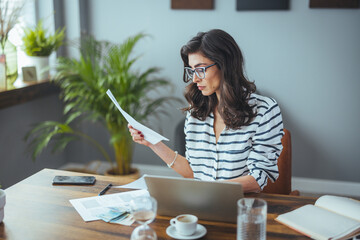 A Caucasian businesswoman in striped attire meticulously reviews documents beside her laptop in a...