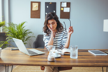 A thoughtful Caucasian woman reads documents at her desk, glasses in hand, with laptop and digital tablet nearby, indicating diligent decision-making in a professional setting.