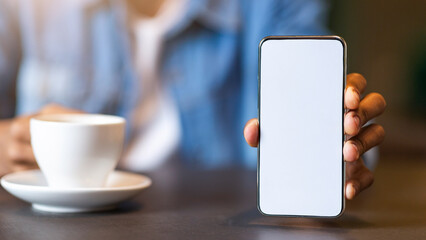 A person sits at a cafe table, holding a smartphone with a blank screen in one hand and a cup of coffee in the other. The atmosphere appears relaxed, suggesting a morning routine or a break.