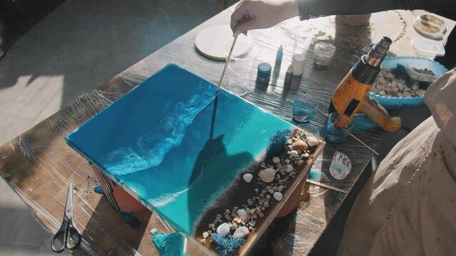 Artist paints the sea using epoxy resin. With the help of a wooden stick, she creates the illusion of sea foam and waves. She works with a blue gradient. Close up. Slow motion