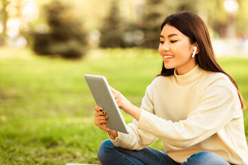 Asian woman sitting on the ground with legs crossed, focused on a tablet held in her hands. She appears engaged and concentrated as she interacts with the device, copy space