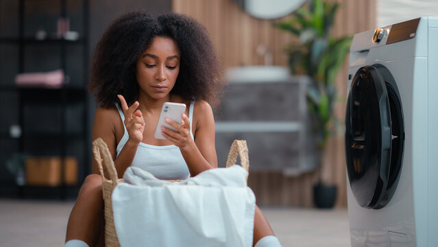 African American woman ethnic girl housewife do housework housekeeping basket with cloth dirty clothes reading instruction using washing machine laundry appliance chatting mobile phone smartphone hold