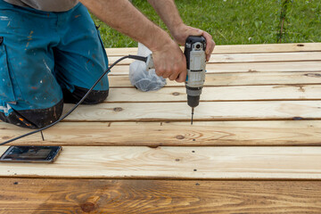 A person wearing blue pants kneels on wooden boards while holding a power drill in their hand to tighten a screw.