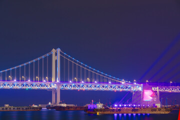 the Gwangan Bridge in Busan, Korea, illuminated at night with vibrant blue and purple lights, reflecting beautifully on the water.