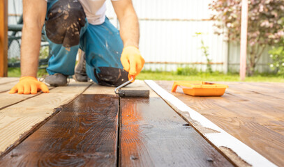 A person is kneeling and using a paint roller to apply a dark wood stain to a wooden deck outside on a sunny summer day.