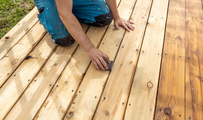A person is kneeling on a wooden deck and sanding the boards with a sanding block.