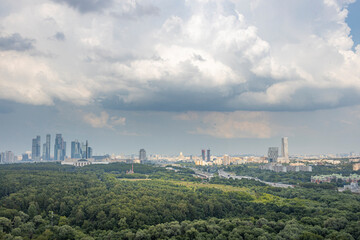 A panoramic view of the Moscow skyline, featuring a dense forest in the foreground and skyscrapers in the distance. The clouds in the sky hint at a potential spring shower.