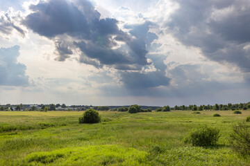 Fototapeta premium A vast green meadow with scattered trees, viewed from a slight elevation, with a dramatic, cloudy sky above.