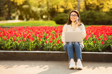 A young Asian woman with glasses is sitting on the edge of a flowerbed filled with blooming tulips while using her laptop. She appears focused and is enjoying the sunny afternoon in the park.